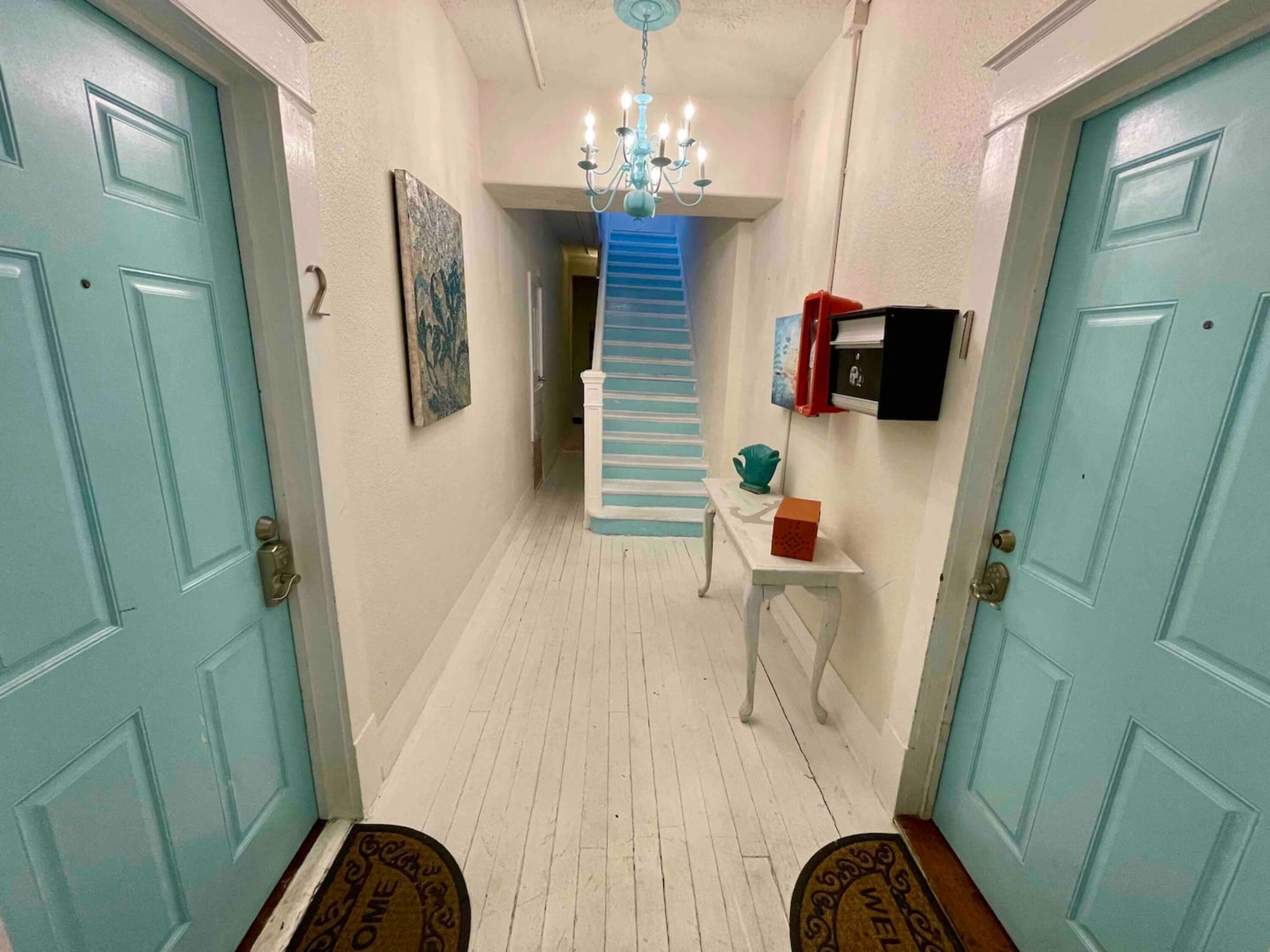 Un pasillo con una escalera, un candelabro, una puerta con felpudo y una mesita con una caja.