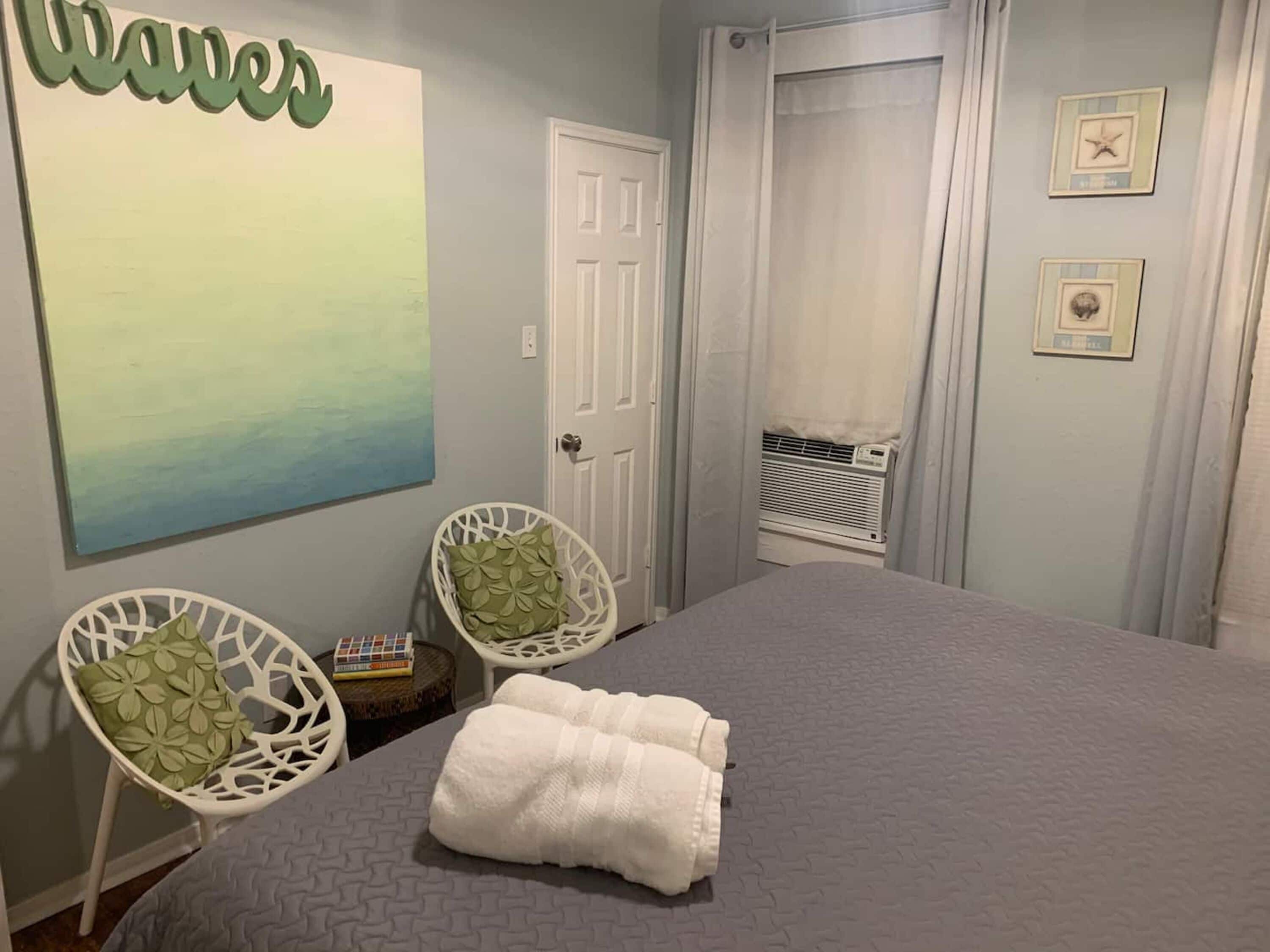 A bedroom with a bed, two white chairs, a wall art piece, a door, and an air conditioning unit.