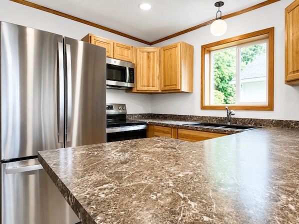 A kitchen with a stainless steel refrigerator, a microwave, an oven, wooden cabinets, a granite countertop, and a window with a view of greenery.