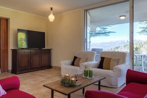 Un salón con un televisor de pantalla plana, dos sofás blancos con cojines, una mesa de centro de madera con velas y vistas a las montañas a través de una puerta de cristal.