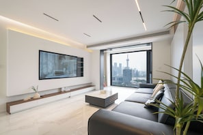 Una sala moderna con un televisor de pantalla plana grande, un sofá, una mesa de centro y vistas a un paisaje urbano a través de un ventanal amplio.