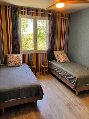 Ein Zimmer mit zwei Betten, einem Fenster mit Vorhängen, einem kleinen Hocker und einer Lampe.