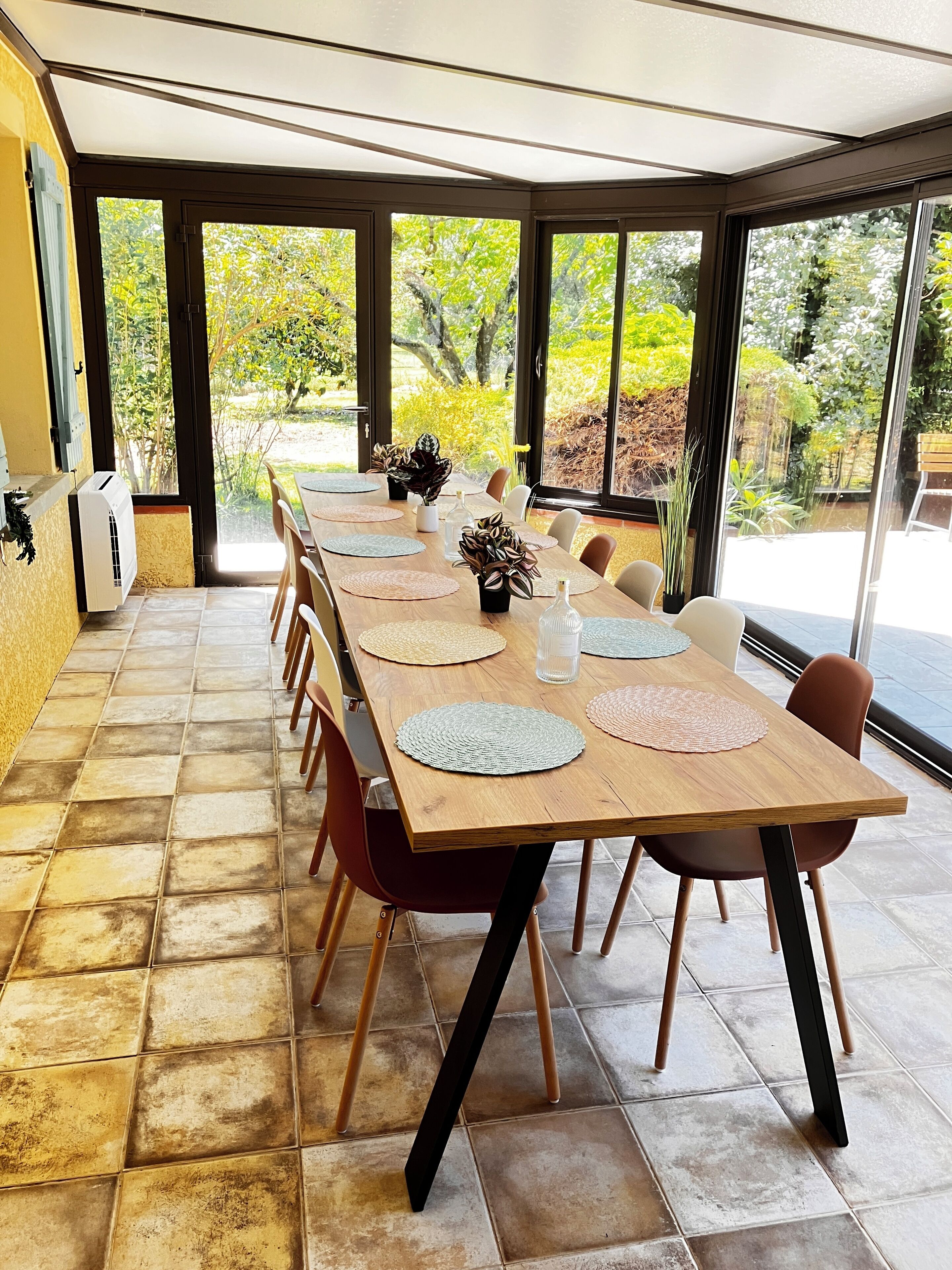 Une longue table à manger entourée de chaises, un ventilateur de plafond et de grandes fenêtres donnant sur le jardin.