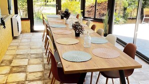 Une longue table à manger entourée de chaises, un ventilateur de plafond et de grandes fenêtres donnant sur le jardin.