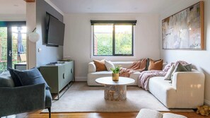 Ein Wohnzimmer mit einer Couch, einem Couchtisch, einem Fernseher und einem Fenster mit Blick auf Grünflächen.