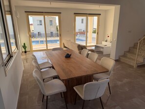 Un'ampia stanza con un grande tavolo di legno e diverse sedie, una scala e vista su una piscina e edifici esterni.