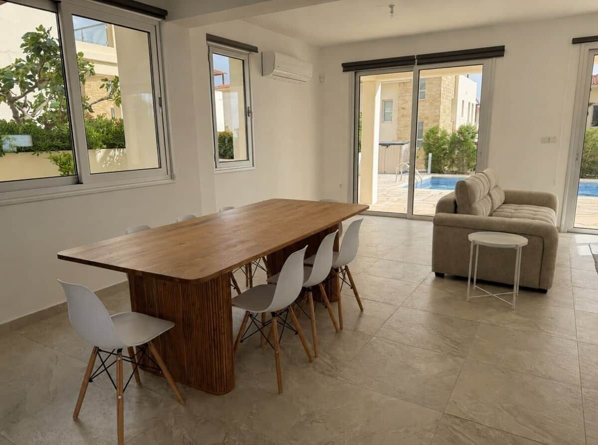 Un salon moderne avec une table à manger en bois, des chaises blanches, un canapé beige et un petit tabouret blanc. De grandes fenêtres offrent une vue sur une piscine et un espace extérieur.