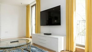 A modern room with a TV mounted on the wall, a glass table with a remote, and a white cabinet. - Modern 1-Bedroom Greenwich Apartment - Close to O2 Arena (London)