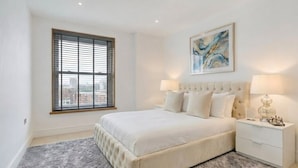 A bedroom with a large bed, two bedside tables, a window with blinds, and a painting on the wall. - Modern 1-Bedroom Greenwich Apartment - Close to O2 Arena (London)