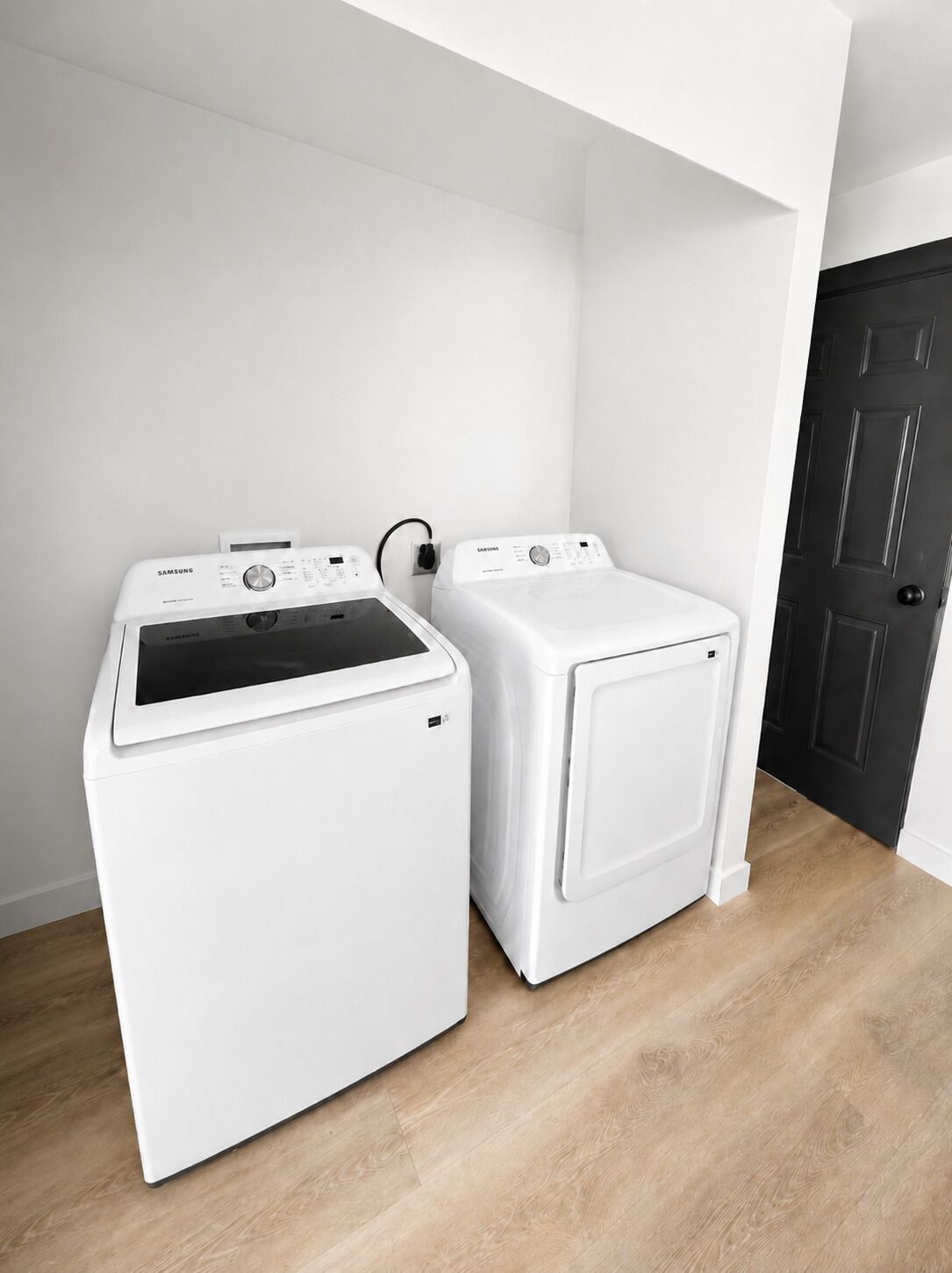 A laundry room with two white washing machines, a black door, and a wooden floor.