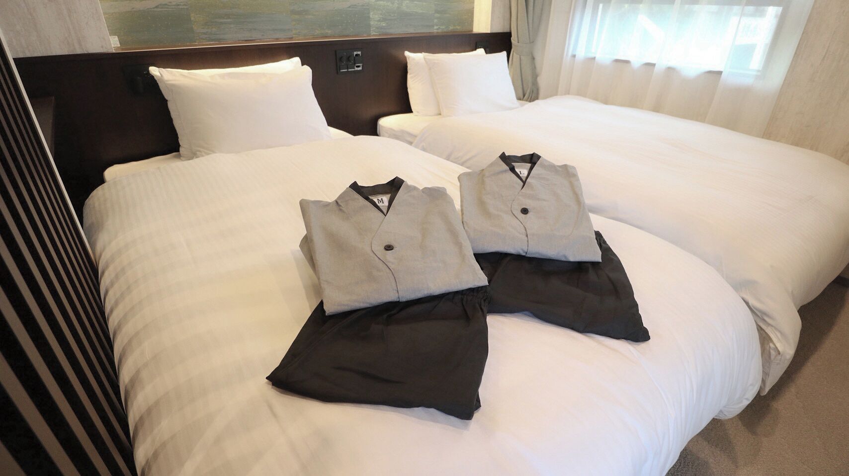 Habitación de hotel con dos camas, cada una con ropa blanca de cama y almohadas. Se observa un conjunto de ropa doblada cuidadosamente sobre las camas.