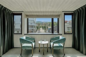 Quarto com duas cadeiras, uma mesa pequena e vista para a cidade através de grandes janelas.