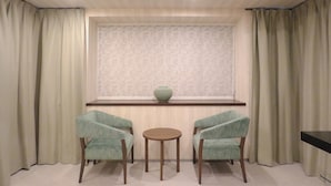 Um quarto com duas cadeiras, uma mesa redonda pequena e uma janela com persiana estampada.