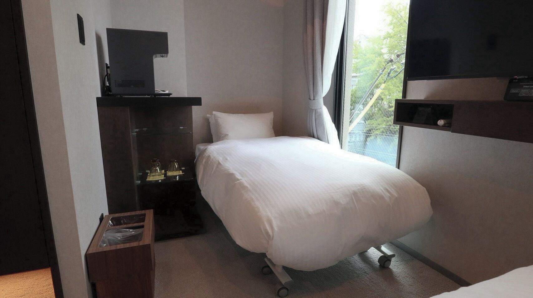 A hotel room with a bed, a television, a desk with a computer, and a window with a view of trees.