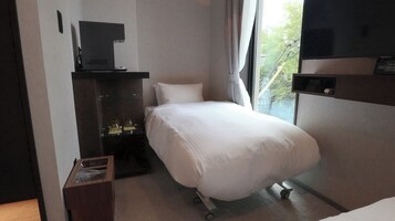 Quarto de hotel com uma cama, televisão, escrivaninha com computador e janela com vista para árvores.