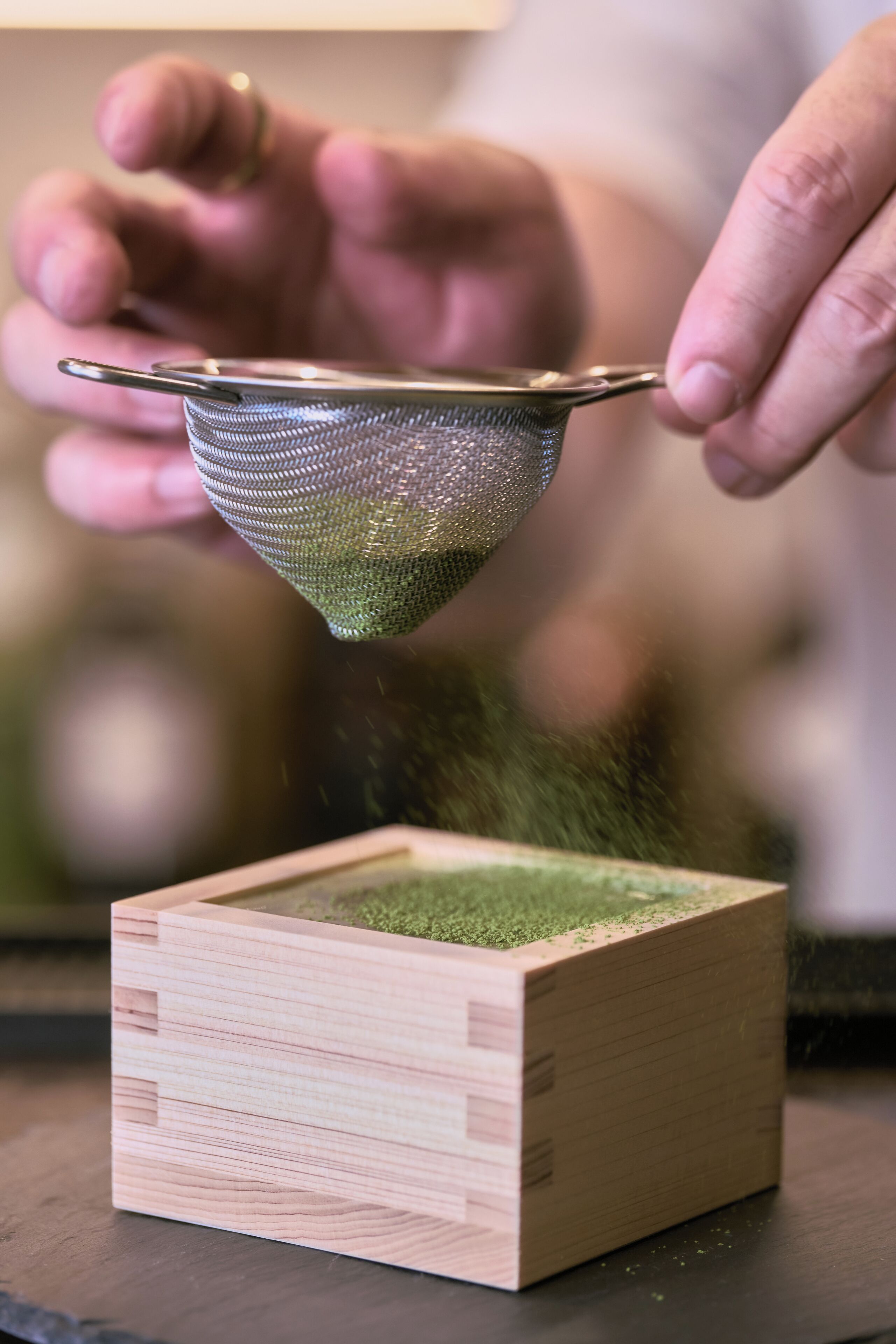 A person using a metal sieve to sift green material from a wooden box.