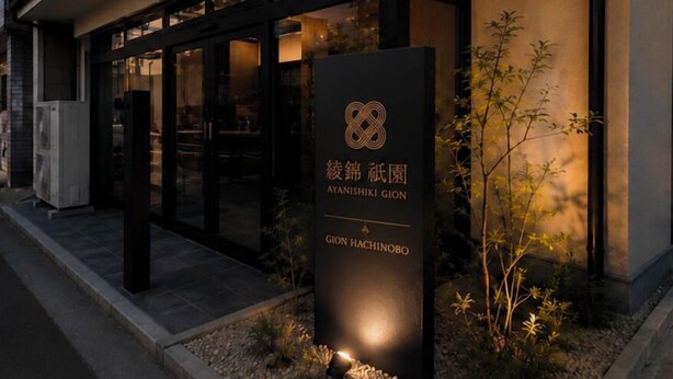 Entrada de um edifício moderno com portas de vidro, uma placa com texto em japonês e uma pequena área ajardinada.