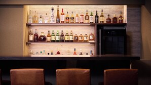 Uma área de bar com várias prateleiras de garrafas de bebidas, uma geladeira e três banquetas de couro.