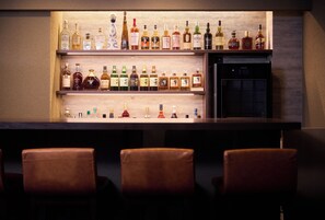 Uma área de bar com várias prateleiras de garrafas de bebidas, uma geladeira e três banquetas de couro.