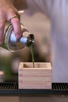 Uma mão despejando um líquido verde de um recipiente metálico para uma caixa de madeira.
