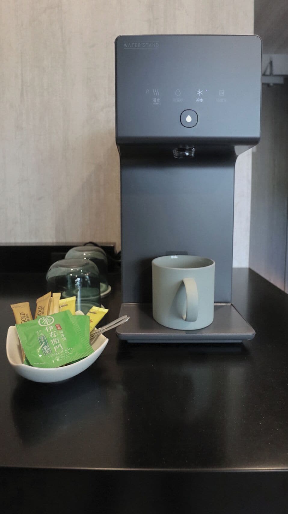 A water dispenser with a cup holder, a bowl of tea bags, and a mug on a countertop.