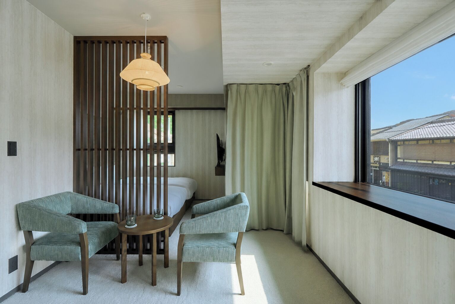 A hotel room with a bed, two armchairs, a small table, a hanging light fixture, a window with a view of buildings, and a wooden partition.