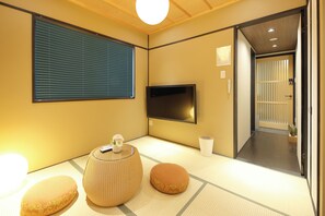 A traditional Japanese room with tatami flooring, a tatami mat table, a low orange cushion, a woven stool, a flat-screen TV, and a sliding door leading to another room. - Ayanishiki Gion (Kyoto)