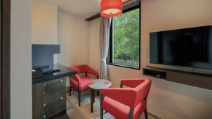 Quarto de hotel com televisão, escrivaninha com um laptop, pequena mesa com duas cadeiras e janela com vista para árvores.