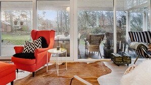 Een modern interieur met een grote glazen deur die leidt naar een terras buiten, een rode fauteuil, een rieten stoel en een gestreepte fauteuil.