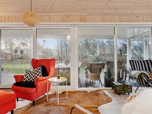 Ein modernes Interieur mit einem großen Glasfenster, das auf eine Terrassensitzgelegenheit führt, ein rotes Sessel, ein Korbstuhl und ein gestreifter Sessel.