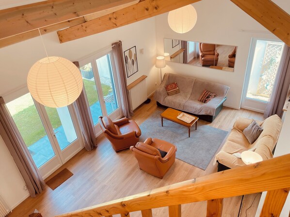 A spacious living area with a sofa, armchairs, and a coffee table. Large windows provide natural light and a view of the outdoors.