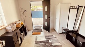 Um corredor com chão de azulejos, uma porta que dá para o exterior, um banco preto, um espelho e um suporte fixado na parede.
