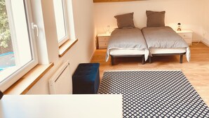 Um quarto com cama, mesas de cabeceira, um banquinho, tapete estampado e uma janela.