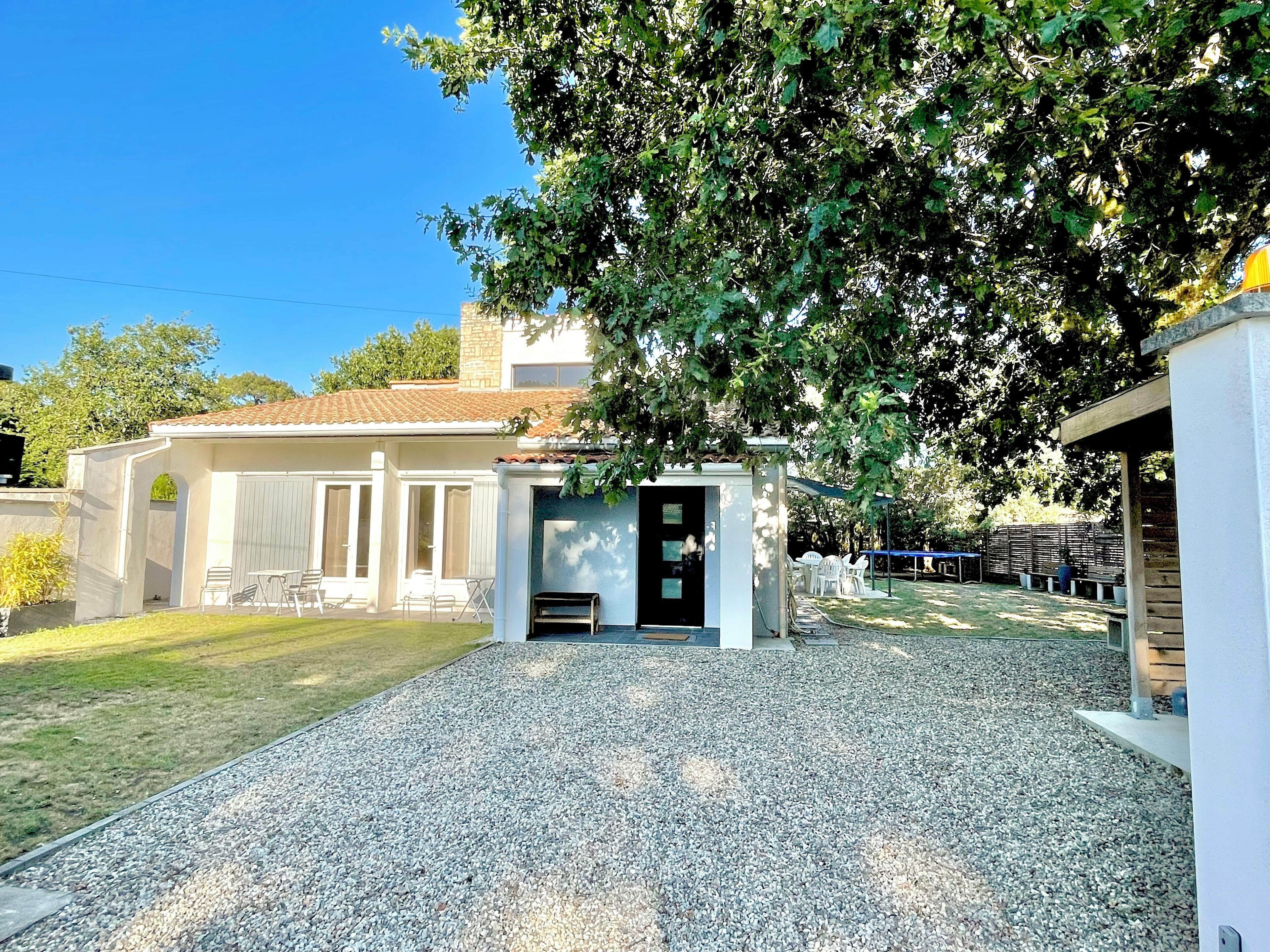 Une maison dotée d’un patio couvert, d’une allée en gravier et d’un grand arbre qui offre de l’ombre.