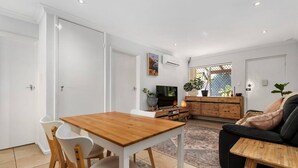 A modern living room with a wooden dining table, white chairs, a black sofa, and a TV. - Edmund Place – Charming Fremantle Retreat (Fremantle)