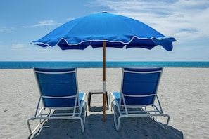 Zwei Strandstühle unter einem blauen Sonnenschirm an einem sandigen Strand, im Hintergrund das Meer.