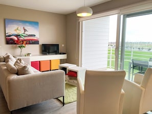 A modern living room with a sofa, a TV, a small table, and a large window with a view of a green field. - Family Duplex Near the Belgian Coast (Nieuwpoort)