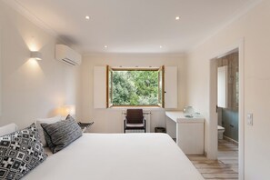 Un dormitorio con cama, escritorio, silla, ventana con vista a árboles y baño visible a través de una puerta abierta.