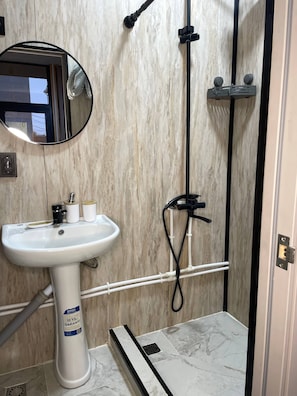 Une salle de bain avec un lavabo sur colonne blanc, un miroir, une douche avec un tuyau noir et un sol carrelé.