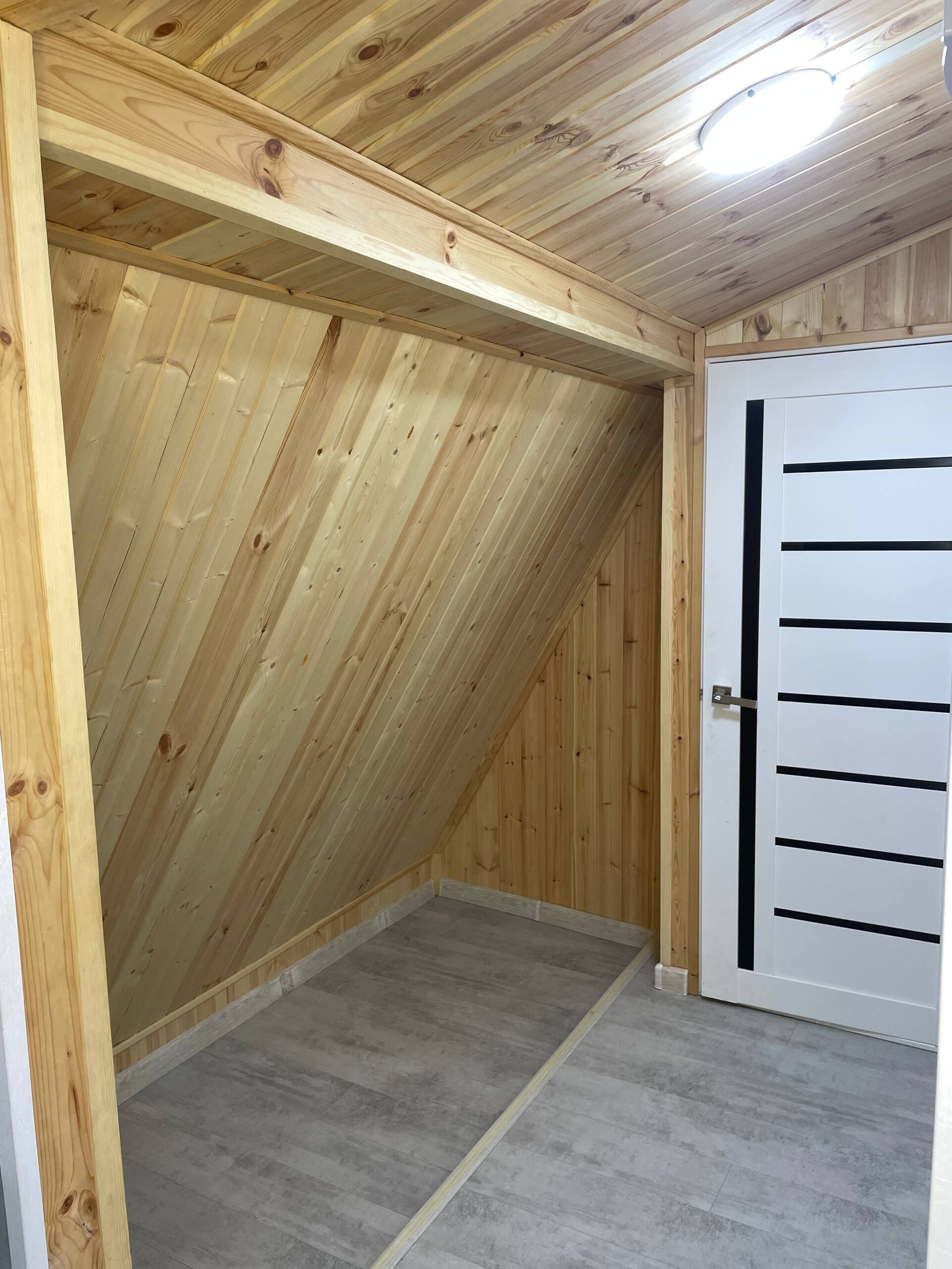 Une pièce mansardée en bois, avec un plafond en pente, une porte à rayures noires horizontales et un luminaire au plafond.