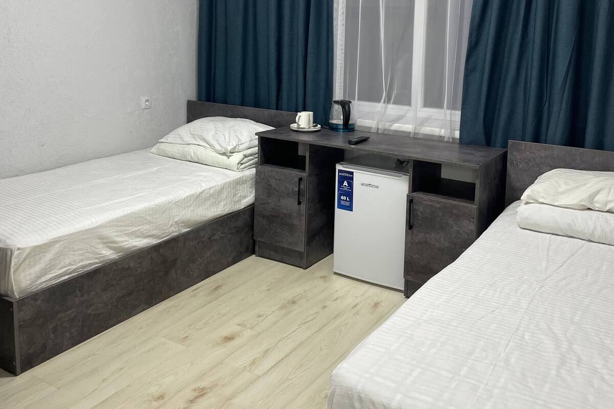 Habitació d'hotel amb dos llits, un escriptori petit, un mini-frigorífic i una finestra amb cortines.