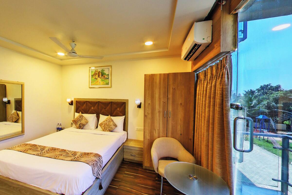 A hotel room with a bed, a chair, a desk, a mirror, a ceiling fan, an air conditioning unit, and a view of the outside.