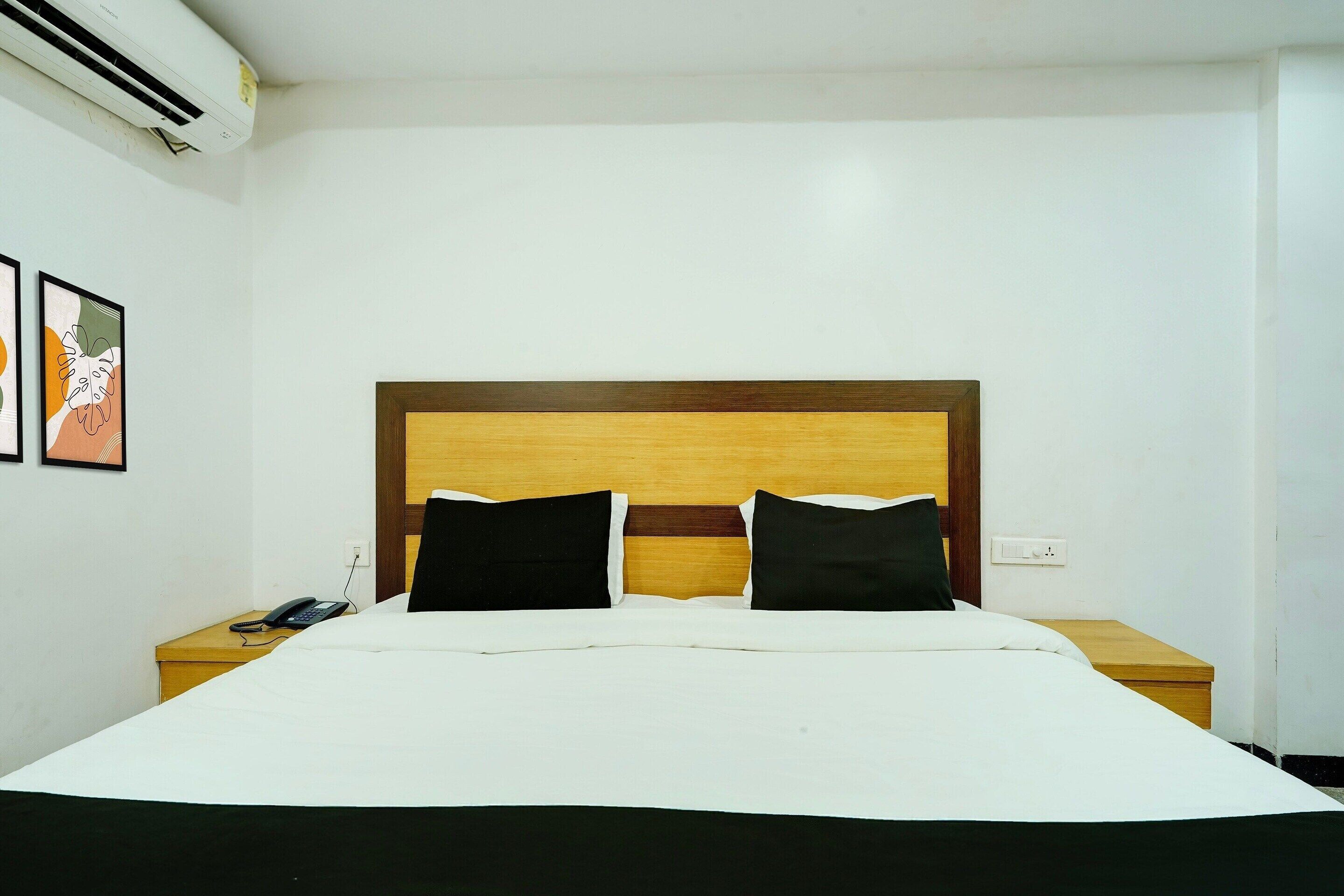 A hotel room with a wooden headboard, white bedding, two black pillows, a bedside phone, and a wall-mounted air conditioner.