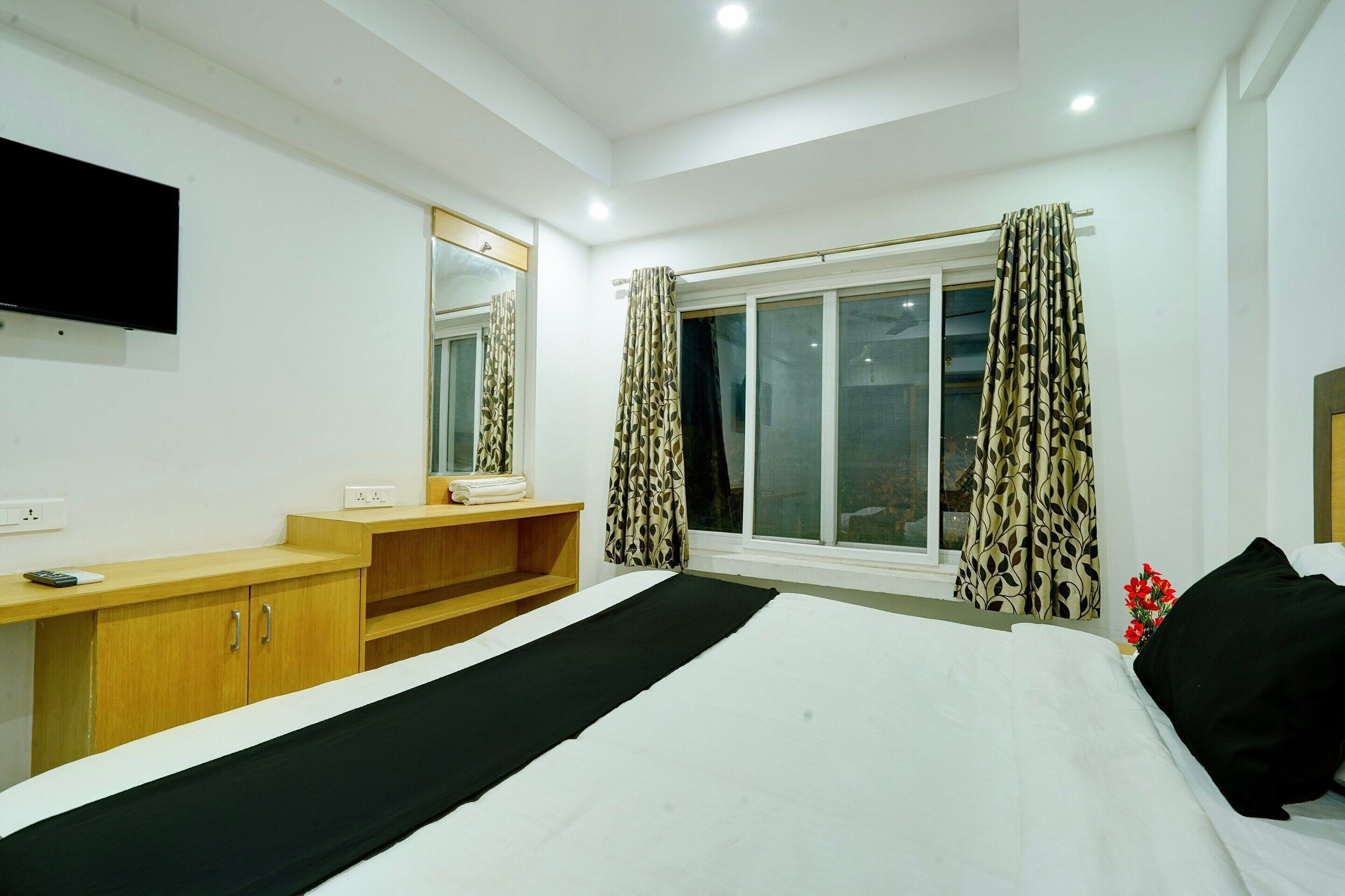 Habitación de hotel con cama, televisor montado en la pared, ventana con cortinas y escritorio de madera con espejo.