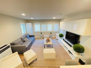 A modern living room with a grey sofa, white ottoman, and a coffee table with fruits. There is a TV on a white cabinet and a small plant on a side table.