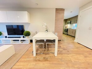 A modern living room with a flat-screen TV, a dining table with chairs, a small potted plant, and a kitchen area in the background.