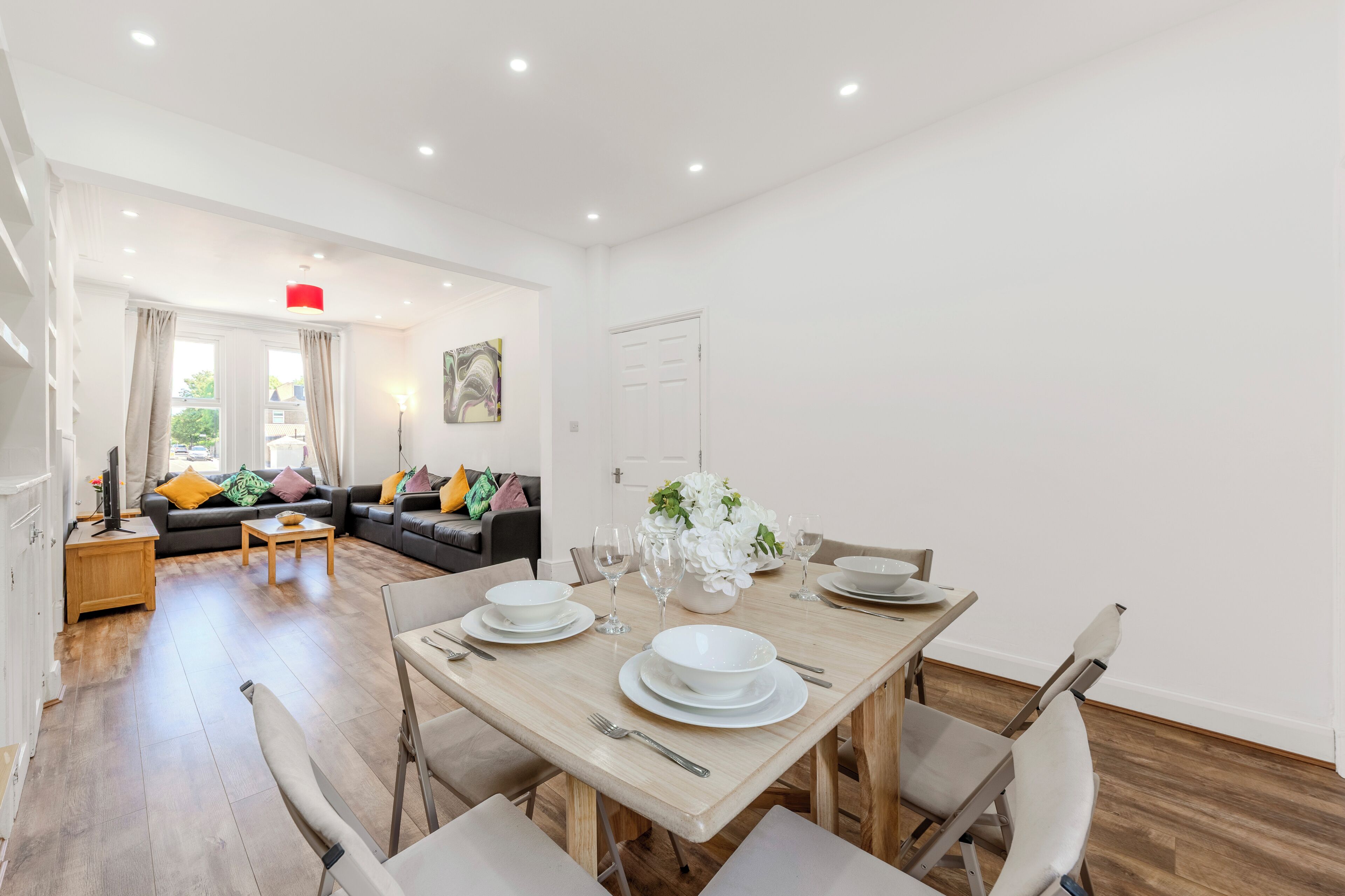 A dining area with a wooden table set for four, a sofa in the background, and a door to the right.