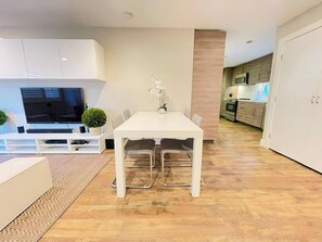 A modern living room with a flat-screen TV, a dining table, and a kitchen in the background.