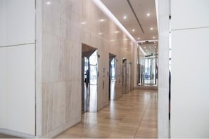 Un pasillo moderno con paredes de mármol y suelo de madera, que conduce a ascensores y una entrada con puerta de vidrio.