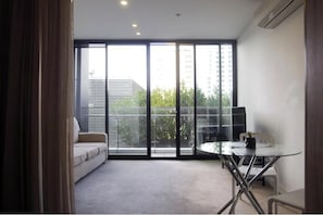 Ein modernes Zimmer mit einer Couch, einem kleinen Tisch und Blick auf Gebäude und Bäume durch große Fenster.